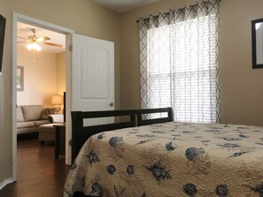 A bedroom with a bed, a door, a ceiling fan, a window with patterned curtains, and a nightstand with a lamp. - "Koastal Escape" 1st Floor 1 BDR-1 Bath/Just steps to the Beach! (Corpus Christi)