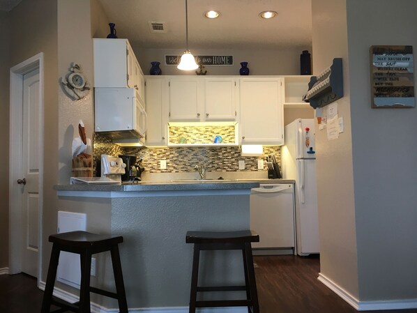 A kitchen with white cabinets, a tiled backsplash, a built-in dishwasher, and two bar stools. - "Koastal Escape" 1st Floor 1 BDR-1 Bath/Just steps to the Beach! (Corpus Christi)
