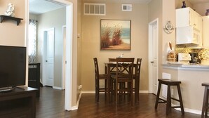 A living room with a TV, a dining area with chairs and a table, and a kitchen with cabinets and a microwave.