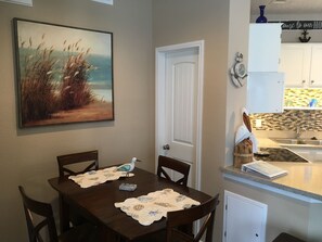 A dining area with a table set for four, a painting on the wall, and a kitchen visible through an open door. - "Koastal Escape" 1st Floor 1 BDR-1 Bath/Just steps to the Beach! (Corpus Christi)