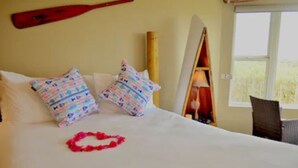 A bed with patterned pillows, a surfboard leaning against the wall, and a red lei on the bed. - Panoramic Windows, Double showers, Fireplace (Moss Landing)