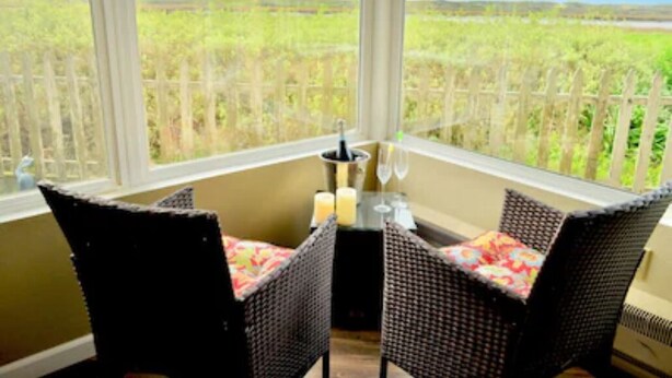 A corner room with wicker chairs and a small table, a window with a view of a fence and greenery. - Panoramic Windows, Double showers, Fireplace (Moss Landing)