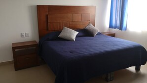 Ein Schlafzimmer mit einem hölzernen Kopfende, einer blauen Bettdecke, zwei Kissen, einem Nachttisch und einem Fenster mit blauen Vorhängen.