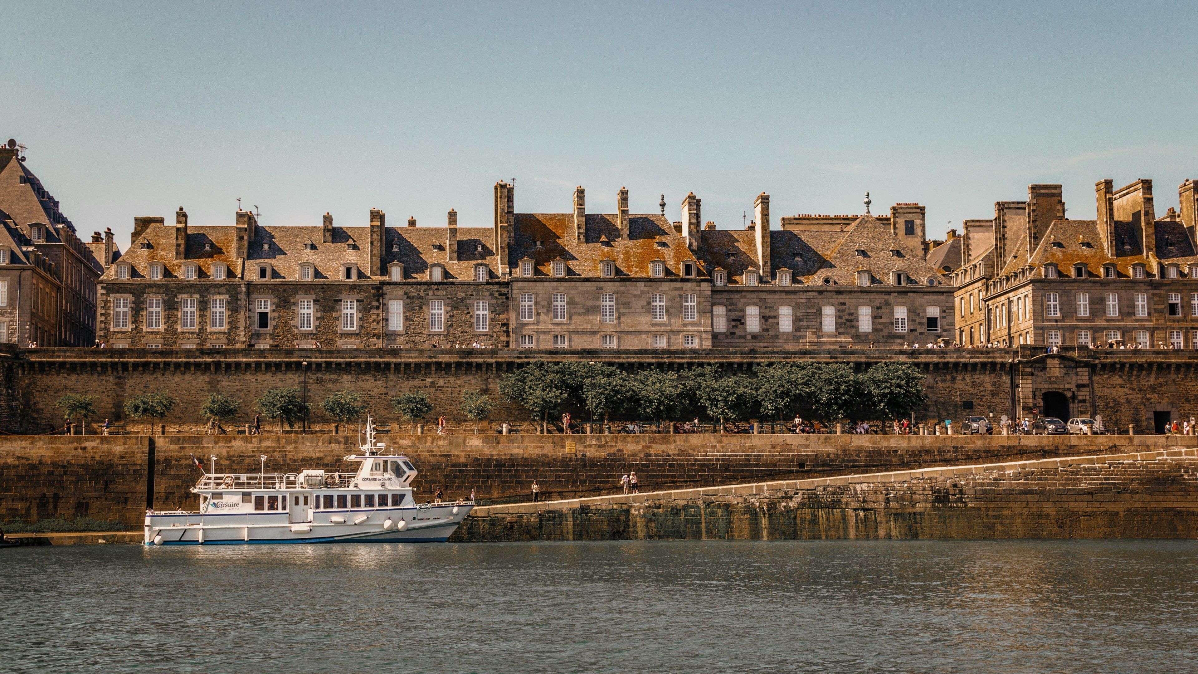 A historic building with a stone facade, a river in the foreground, and a boat docked at the riverbank.
