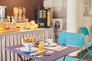Una barra de desayuno con máquinas de café, una selección de pasteles y un área de estar con una silla y una mesa.