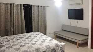 Een slaapkamer met een bed, een grijze bank, een flatscreen televisie aan de muur en een plafondventilator.