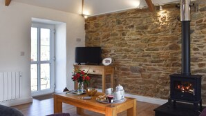 A living room with a stone wall, a fireplace, a television, and a dining table with a vase of flowers. - 3 bedroom accommodation in Mount Hawke, near St Agnes (Mount Hawke, near St Agnes)