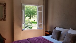 Een slaapkamer met een bed, een raam met vrij uitzicht op groen en een nachtkastje met een fles.