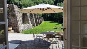 Een terras buiten met stenen muren, een gazon en een tafel met stoelen onder een grote parasol.