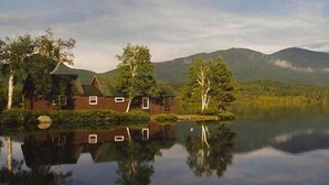 Une maison au bord du lac, dont l’image se reflète dans l’eau, entourée d’arbres et de montagnes.