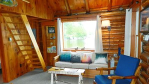 L’intérieur d’une cabane, avec un escalier en bois, un canapé agrémenté de coussins colorés, un fauteuil bleu et une fenêtre donnant sur un lac.