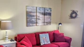 Un sofá rojo con una almohada decorativa, una lámpara sobre una mesita de noche y dos cuadros colgados en la pared.