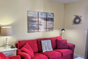 Un canapé rouge agrémenté d’un seul coussin décoratif, une lampe sur une table de chevet et deux tableaux accrochés au mur.