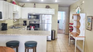 A kitchen with white cabinets, a microwave, stove, and refrigerator. There are two stools and a shelf with red items. - Dog-Friendly·Gulf Views· Steps to White Sand Beach (Port Saint Joe)