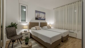 Een slaapkamer met twee bedden, een nachtkastje, een stoel, een kleine tafel met een lamp en een raam met gordijnen.