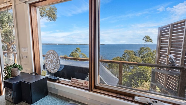 A room with a large window overlooking a body of water, a small table with a plant, a decorative item, and a 'Thank you for not smoking' sign. - Lakescapes Cottage: Relax & enjoy 180 degrees, uninterrupted water views (Metung)