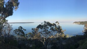A coastal landscape with a calm sea, dense trees, and a distant shoreline with buildings. - Lakescapes Cottage: Relax & enjoy 180 degrees, uninterrupted water views (Metung)