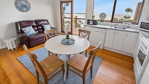 A cozy living area with a round table, wooden chairs, a brown leather sofa, and a view of the sea through large windows. - Lakescapes Cottage: Relax & enjoy 180 degrees, uninterrupted water views (Metung)