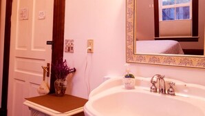Ein Badezimmer mit einem weißen Waschbecken, einem Spiegel mit floraler Umrandung und einem Blumenvasen mit lila Blumen auf der Ablagefläche.