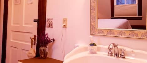 Un baño con un lavabo blanco, un espejo con marco floral y un florero con flores moradas sobre la encimera.
