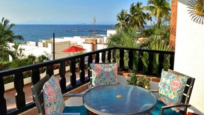 Un balcón con una mesa y sillas de vidrio, con vista a la costa, palmeras y barcos.