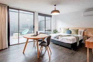 A modern hotel room with a large bed, a round dining table, and a view through a sliding glass door.