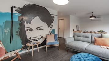 A modern living room with a large abstract painting, a grey sofa, a blue ottoman, and a wooden coffee table.