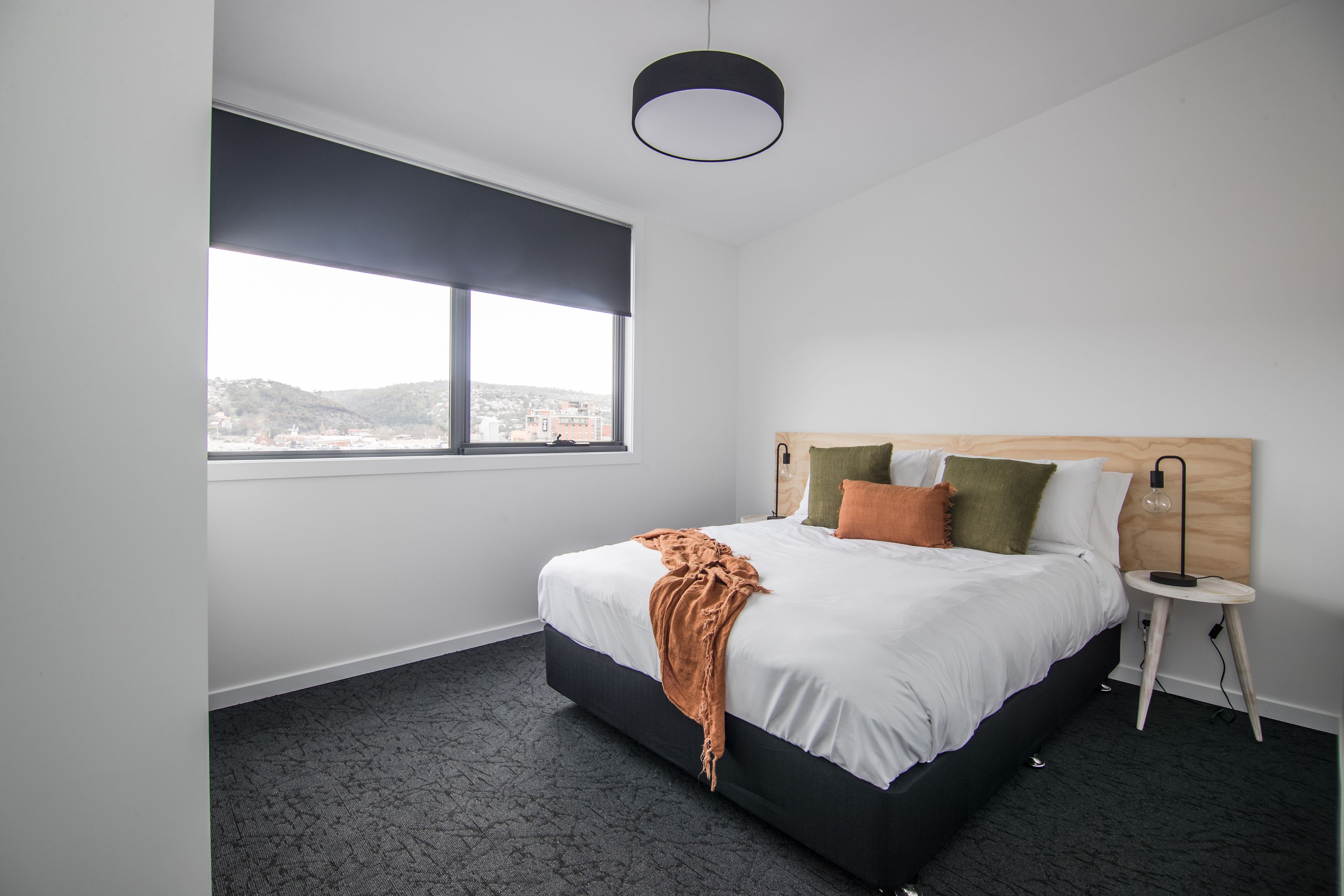 A modern hotel room with a large bed, a side table, a lamp, and a view of the city through a large window.