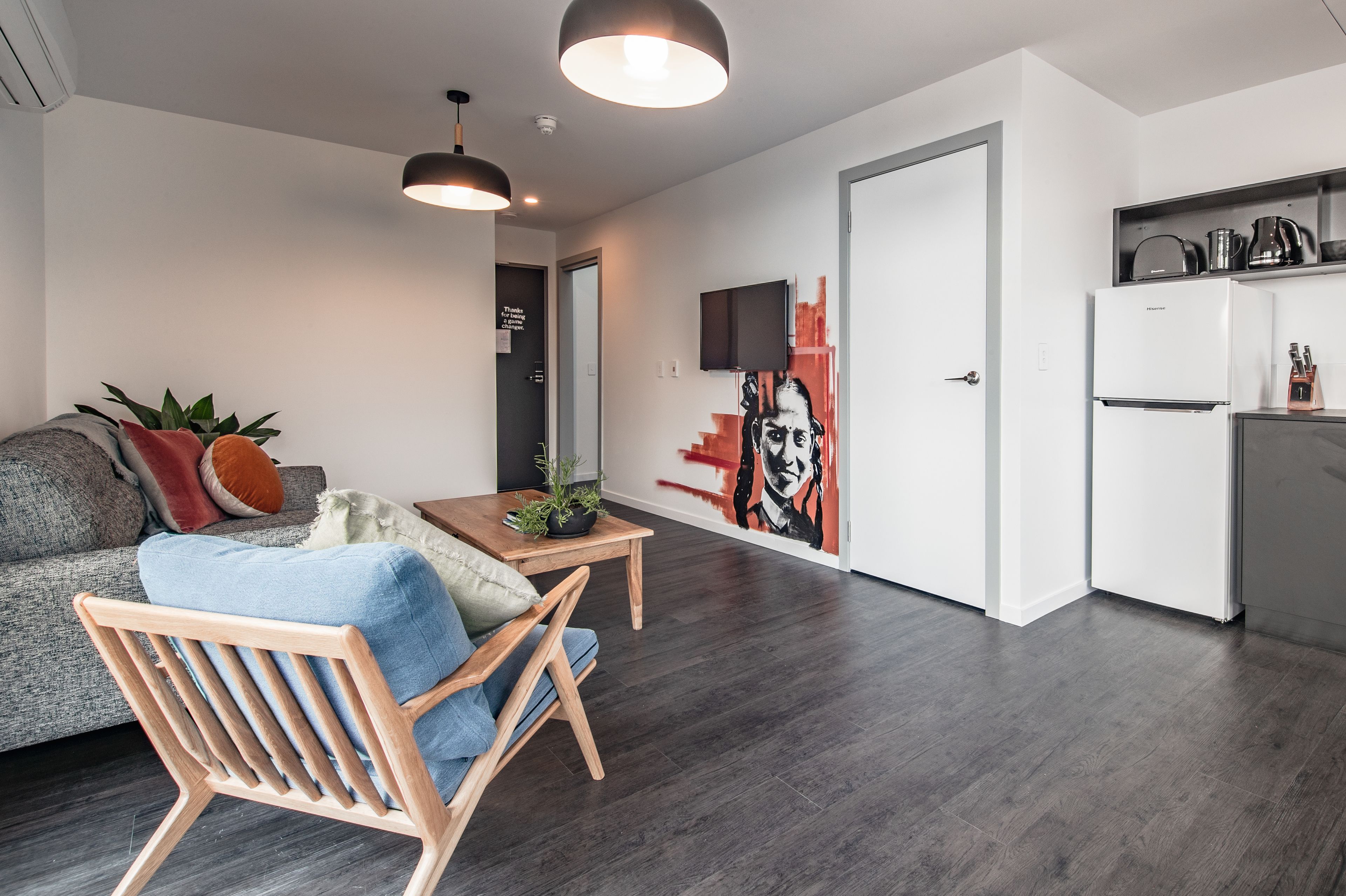 A modern living room with a sofa, a wooden chair, a small table with a plant, and a kitchen area with a refrigerator and a TV.