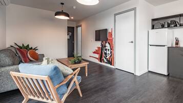 Ein modernes Wohnzimmer mit einer Couch, einem Holzstuhl, einem kleinen Tisch mit einer Pflanze sowie einer Küchenzeile mit Kühlschrank und Fernseher.