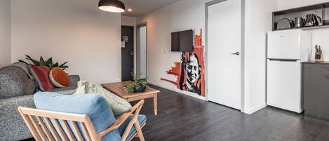 A modern living room with a sofa, a wooden chair, a small table with a plant, and a kitchen area with a refrigerator and a TV.