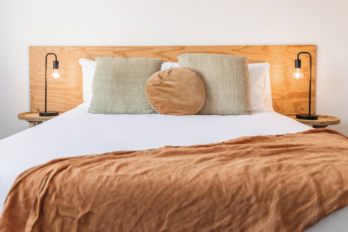 Una cama bien hecha con una manta marrón, dos mesitas de noche con lámparas y un cabecero de madera.