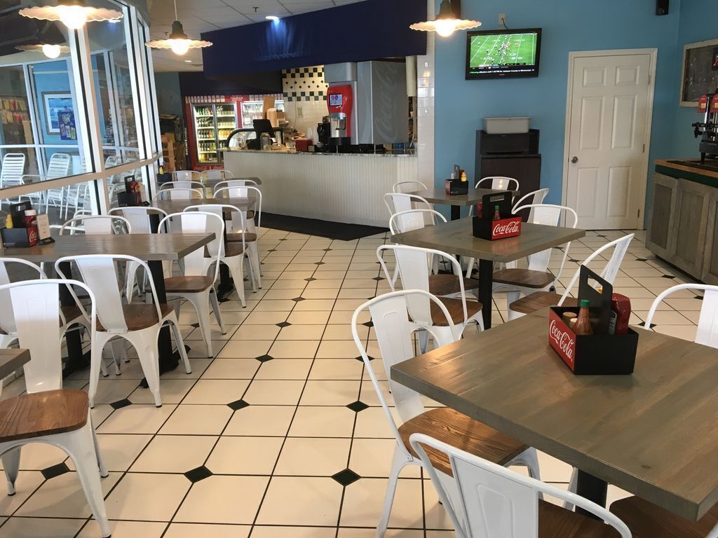 Interior de restaurante com cadeiras brancas, mesas de madeira, chão de azulejos, balcão com refrigerador de bebidas e uma televisão na parede.