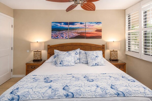 A bedroom with a bed, two bedside tables, a ceiling fan, a window with blinds, and a wall-mounted painting.
