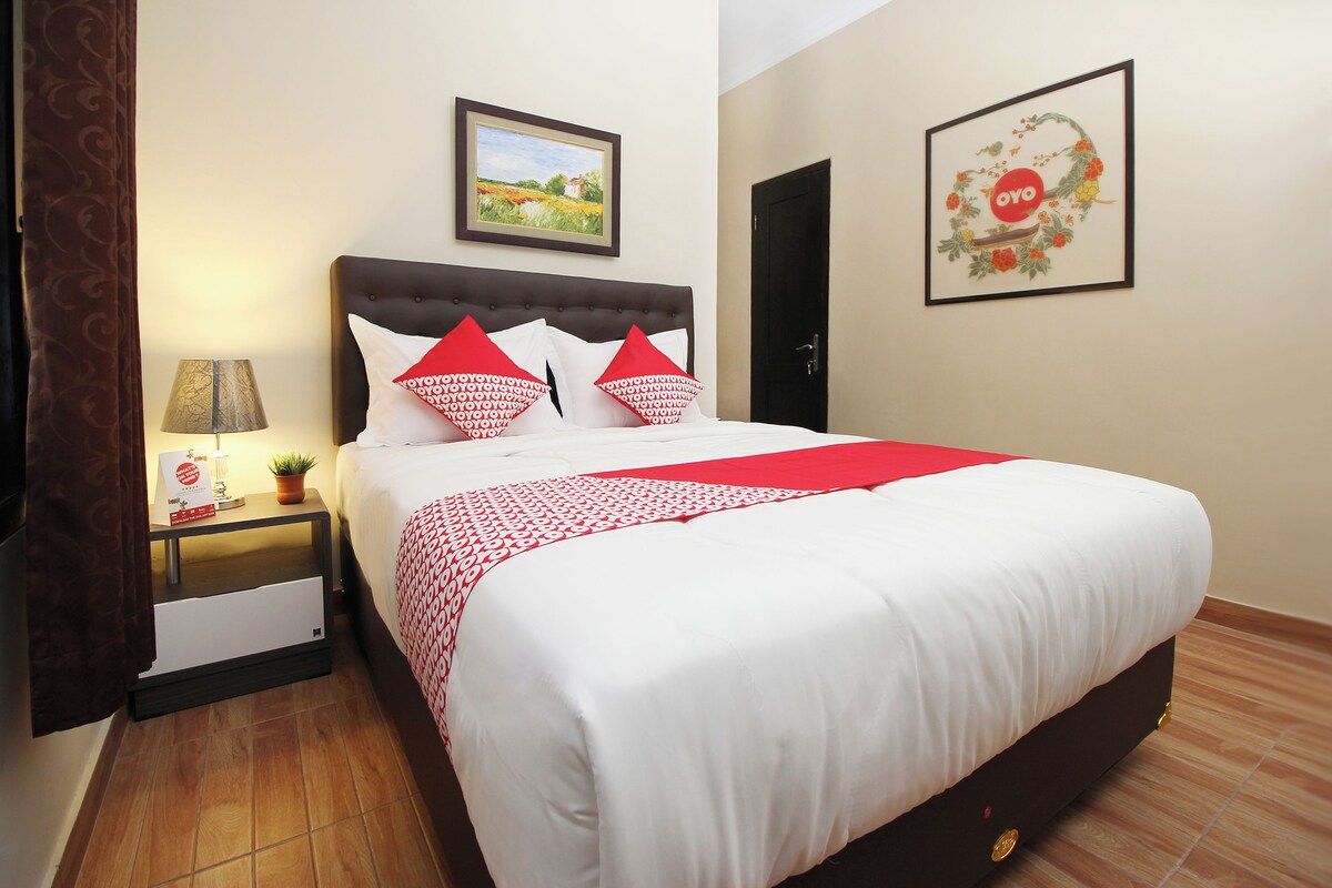A neatly made bed with white and red bedding, a nightstand with a lamp and books, and two framed pictures on the wall.