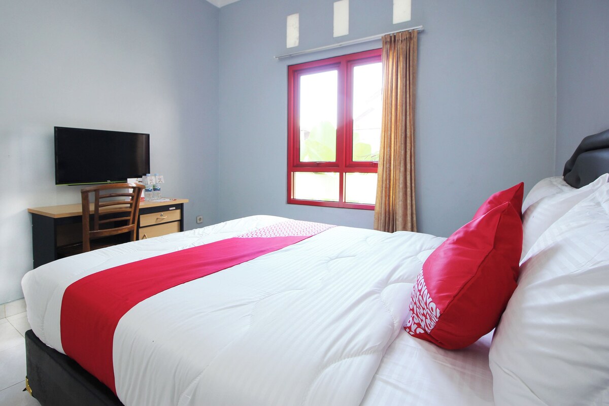 A neatly made bed with white and red bedding, a television on a wooden stand, and a window with curtains.