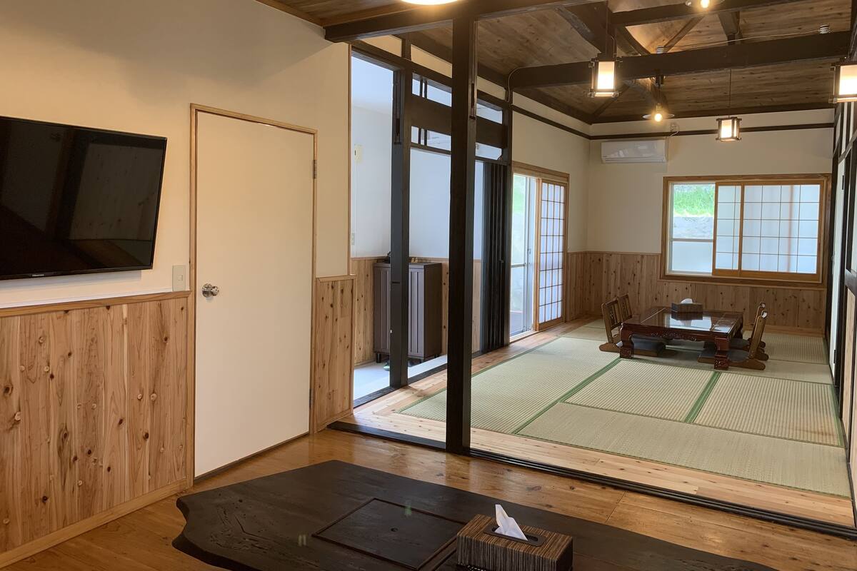 A traditional Japanese room with tatami flooring, a tatami mat table, sliding doors, and a television mounted on the wall.