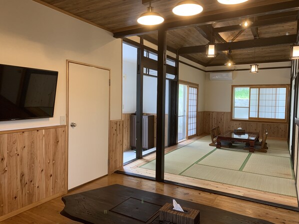 A traditional Japanese room with tatami flooring, a tatami mat table, sliding doors, and a television mounted on the wall.