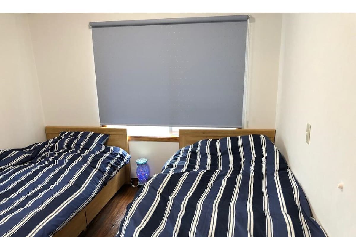 A room with two single beds, a window with a blind, and a small blue vase on the floor.