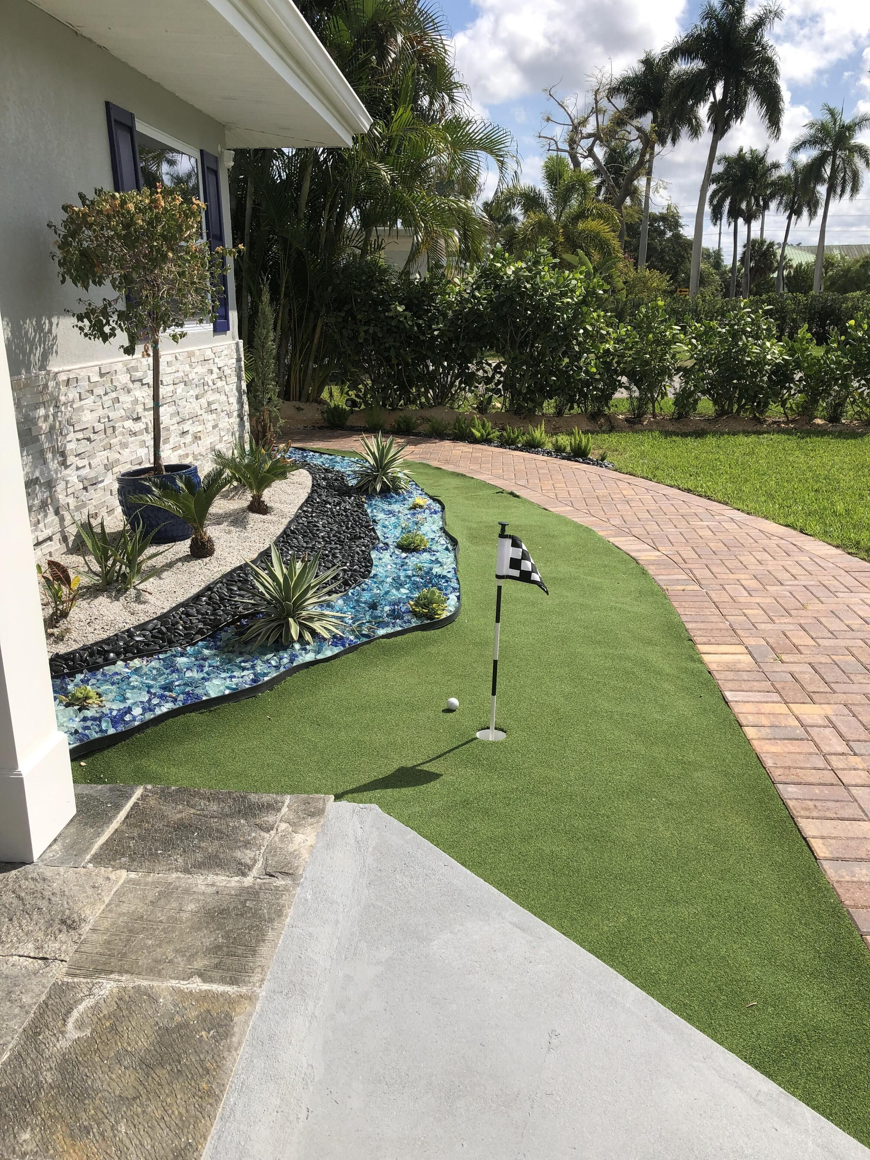 A well-maintained lawn with a golf hole and flagstick, surrounded by a stone wall and tropical plants.