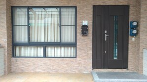 Eine Gebäudefassade mit einem großen Fenster, einer dunklen Tür und zwei Stromzählern auf der rechten Seite.