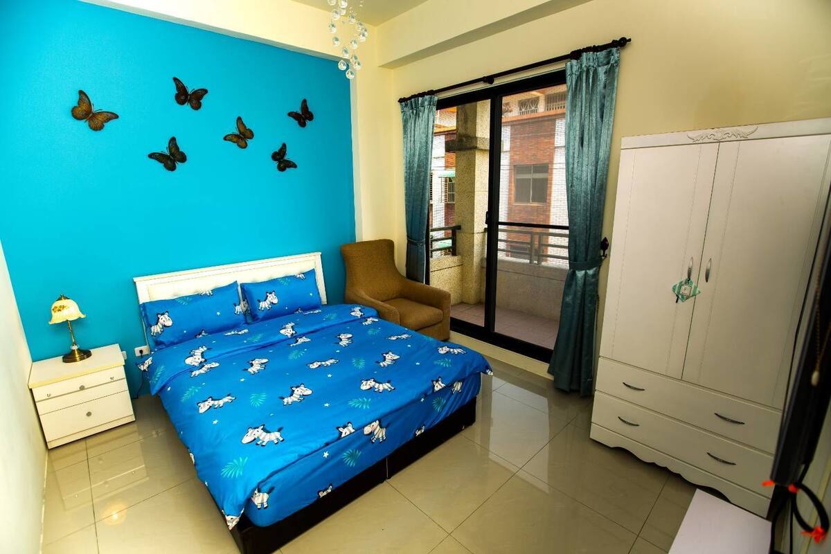 Un dormitorio con colcha azul, una mesita de noche con lámpara, una silla, un armario y un balcón con cortinas.