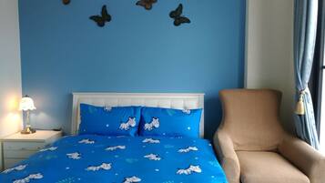 A bedroom with a blue bedspread featuring animal prints, a white headboard, a beige armchair, a nightstand with a lamp, and a window with curtains.