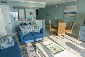 Un salon avec un canapé bleu, des fauteuils en osier, une table basse blanche et un coin cuisine décoré d’une étoile de mer.