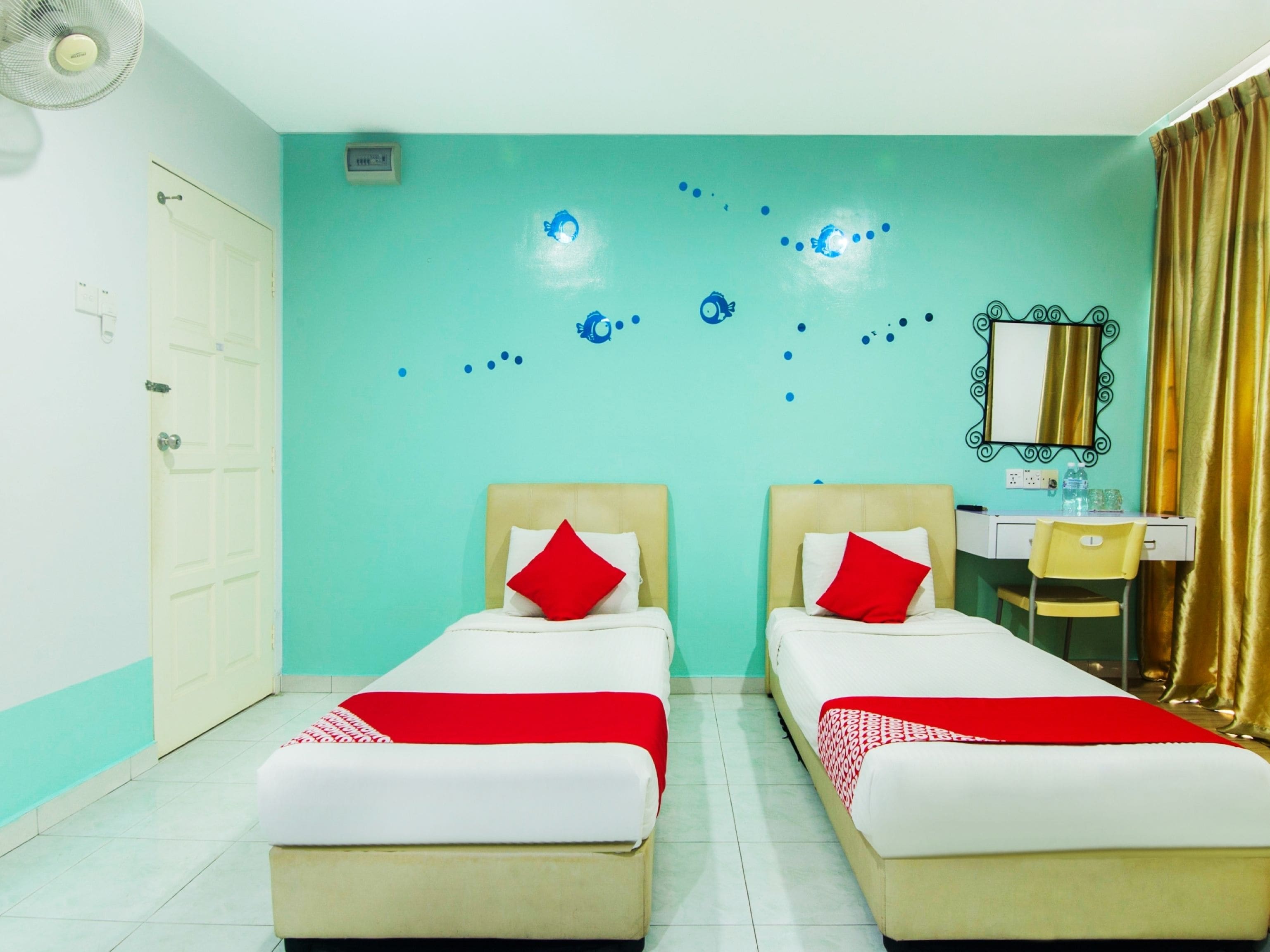 A hotel room with two beds, a desk, a chair, a mirror, and a wall with blue decorative elements.