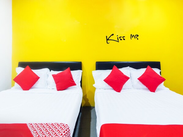 Dos camas con ropa blanca y almohadas rojas frente a una pared amarilla, con la frase 'Kiss Me' escrita encima.