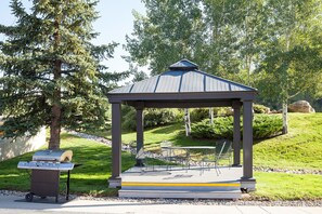 A gazebo with a metal roof and a bench inside, surrounded by greenery and a paved area.