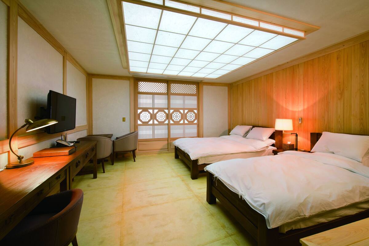 A hotel room with two beds, a desk with a lamp, a television, and a traditional sliding door.