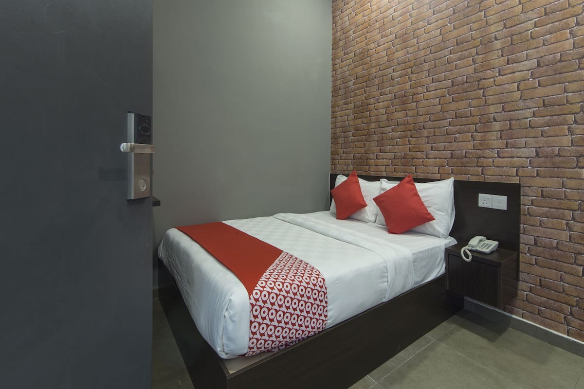 Une chambre d’hôtel avec un mur décoré d’un motif en briques, un lit avec une literie blanche et des oreillers rouges, un téléphone sur la table de chevet et une table de chevet de couleur sombre.