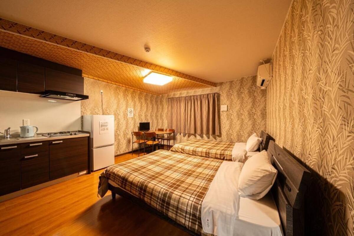 A compact hotel room with a kitchenette, two beds, a desk, and a patterned wall.